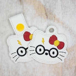 Könnte beinhalten: Zwei weiße Leder-Schlüsselanhänger mit gestickten Bildern einer Comic-Katze mit Brille und einer roten Schleife. Die Katze hat einen Blitz auf der Stirn.