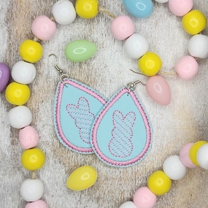 May include: A pair of pastel blue teardrop-shaped earrings with pink stitching. Each earring features an embroidered bunny silhouette. The earrings are displayed against a background of colorful wooden beads.