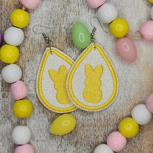 May include: A pair of white teardrop-shaped earrings with a yellow glitter finish. Each earring features a yellow embroidered bunny silhouette.