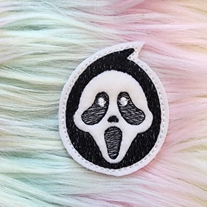 May include: A white ghost face embroidered patch with black stitching on a black circle. The patch is on a rainbow colored fuzzy background.