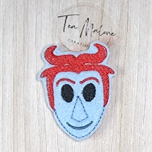 May include: A blue felt patch with an embroidered face of a character with red hair and horns. The character has a mischievous smile and black eyes.