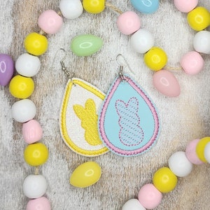 May include: A pair of pastel blue and white teardrop-shaped earrings with embroidered bunny silhouettes. The earrings are hanging from silver hooks.