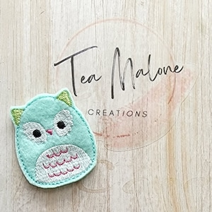 May include: A light blue felt owl applique with green eyes and a white and pink patterned belly. The owl is embroidered with a pink beak and white feathers.