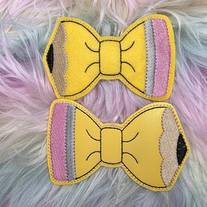 May include: Two yellow bow hair clips, each designed to resemble a pencil. The pencil-shaped bows have black outlines, with a pink, gray, and silver section on one end and a brown and black section on the other.