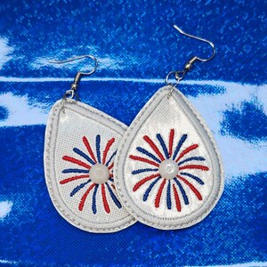 May include: A pair of white teardrop-shaped earrings with red, white, and blue firework designs. The earrings have silver hooks.
