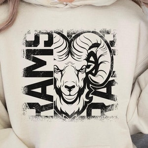 May include: Cream-colored hoodie with a black graphic of a ram's head and the word "RAMS" vertically on the left and "DAZE" vertically on the right. The hoodie has a front pocket and a drawstring hood.