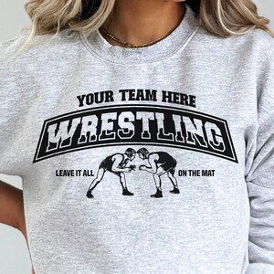 Puede incluir: Sudadera gris con un gr&aacute;fico negro de dos luchadores en una estera. El gr&aacute;fico dice "Your Team Here Wrestling Leave It All On The Mat."