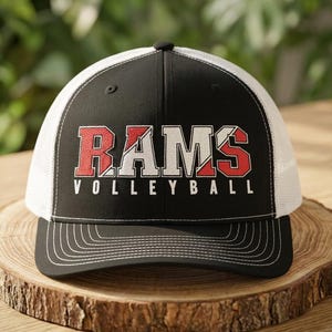 May include: A black and white trucker hat featuring a structured front panel with the text RAMS in a split red and white block font, with the word VOLLEYBALL printed in white underneath. The cap has a black brim with white stitching detail.