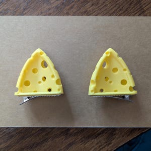 May include: Two yellow cheese slice hair clips with holes, attached to silver metal clips. The clips are arranged on a brown rectangular surface, likely for display or sale. The cheese slices have a textured appearance.