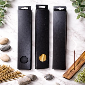 May include: Three black rectangular boxes with a small hole and a hanging tab. One box is open, revealing incense sticks. An incense stick is lit, emitting smoke. The scene includes a wooden incense holder, stones, and greenery.