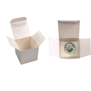 May include: Two small, white cardboard boxes. One box is open, displaying its interior. The other box is open, revealing a silver coin inside. The coin features a profile of a person's head.