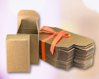May include: Cardboard gift boxes in various stages of assembly. One box is closed, another is open, and a stack of boxes is tied with an orange ribbon. The boxes are a natural brown color, suitable for packaging small items.