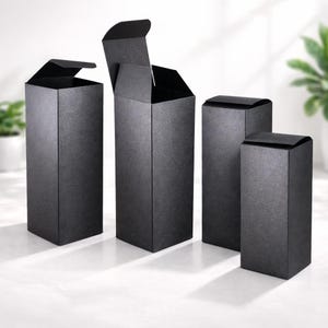 Puede incluir: Cuatro cajas rectangulares negras con superficies texturizadas están dispuestas sobre una superficie blanca. Una caja está abierta, revelando el interior, mientras que las otras están cerradas. Las cajas son altas y estrechas, con un diseño simple y elegante.