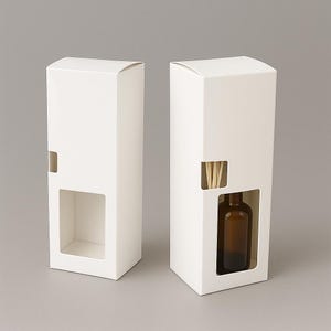 May include: Two white cardboard boxes with cut-out windows. One box is empty, the other contains a brown glass bottle and wooden reeds. The boxes are designed for aromatherapy diffusers or similar products.