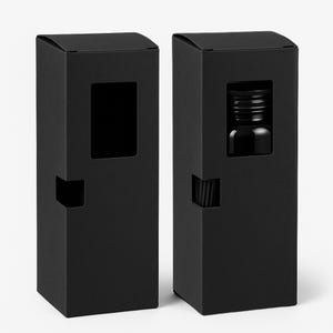 Puede incluir: Dos cajas de cartón negras con una ventana recortada. Una caja contiene una pequeña botella de vidrio. La botella tiene una tapa negra.