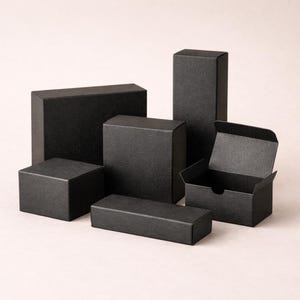 May include: A collection of various sized black gift boxes. The boxes are rectangular and square, with a textured surface. One box is open, revealing the interior. The boxes are arranged on a light pink background.