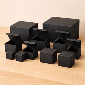 May include: A collection of black square boxes of varying sizes, some with lids open, arranged on a light-colored wooden surface. The boxes are all matte black and appear to be made of cardboard or a similar material.