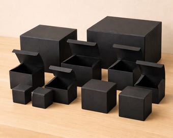 May include: A collection of black square boxes of varying sizes, some with lids open, arranged on a light-colored wooden surface. The boxes are all matte black and appear to be made of cardboard or a similar material.