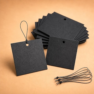 May include: A stack of square black paper tags with a matte finish, each featuring a single circular hole at the top. One tag is attached to a thin black elastic loop, and several additional black elastic loops are arranged separately nearby.