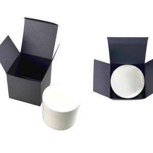 May include: A white round container with a black lid inside a black box with the lid open. The box is on a white surface. There is another black box with the lid open and a white round container inside.