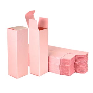 Puede incluir: Se muestran varias cajas rectangulares rosas, algunas ensambladas y otras planas. Las cajas están hechas de un material de papel rosa claro. Las cajas ensambladas tienen una forma alta y delgada con una solapa superior. También se ve una pila de cajas planas.