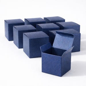 Puede incluir: Nueve cajas de regalo cuadradas azul marino, una abierta, dispuestas sobre una superficie blanca. Las cajas tienen un aspecto texturizado y un diseño simple y elegante. Ideales para pequeños regalos.