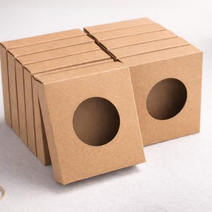 May include: A stack of kraft paper boxes with a circular cutout on the front. The boxes are a light brown color and are arranged in a neat stack. The boxes are likely for packaging small items.