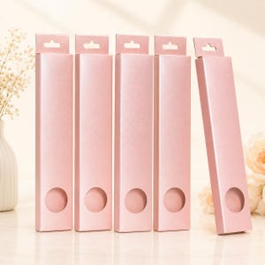 May include: Several pink rectangular boxes with a circular cutout and a small hole at the top for hanging. The boxes are arranged upright, with one leaning slightly to the side. The background is a soft, neutral color.