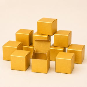 May include: A collection of gold-colored, square gift boxes, some stacked and others arranged around an open box. The boxes have a metallic sheen and are empty. The boxes are suitable for small gifts or favors.