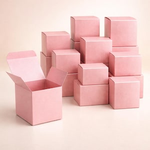 Puede incluir: Una colección de cajas de regalo cuadradas rosas, algunas apiladas y otras abiertas. Las cajas son de un color rosa suave y sólido y parecen ser de cartón. Una caja está abierta, revelando el interior.
