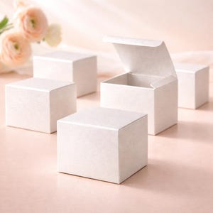 May include: A collection of small, square, white gift boxes, some open, are displayed on a pale pink surface. The boxes have a textured finish, possibly paper or card. Soft pink flowers are visible in the background, adding a delicate touch.