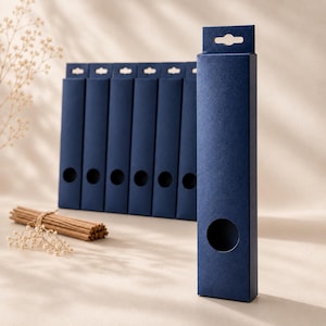 Puede incluir: Cajas rectangulares azul marino con un recorte circular y una pestaña para colgar. Un manojo de palitos marrones atados con hilo y ramitas de flores blancas están en primer plano. Las cajas están dispuestas en fila.