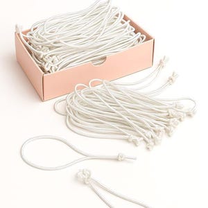 May include: A box filled with white elastic cord with knots at the ends. The cord is arranged in a loose pile. There are two separate pieces of cord in the foreground, each with a knot at one end.