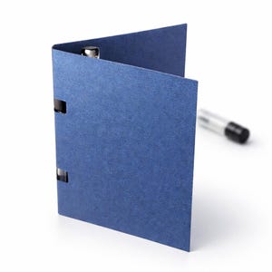 May include: A blue card holder with silver metal accents. The card holder is open, revealing the interior. A pen is slightly out of focus in the background. The card holder is made of a textured material.