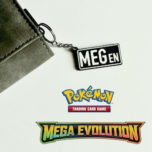 May include: A grey wallet with a silver keyring attached to a black and white rectangular tag that reads "MEGEN". Below the tag are the logos for "Pokémon Trading Card Game" and "Mega Evolution".