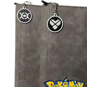 May include: Two gray Pokemon binder tags with metal rings. The left tag features a black and white symbol with a circle and two crescent moons. The right tag features a black and white symbol with a heart shape and two wings. The tags are attached to a gray binder with the text "Pokemon XY The Series" printed on the bottom right corner.