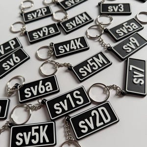 May include: A collection of rectangular black keychains with white borders and white alphanumeric text including sv1V, sv10, sv7a, sv2P, sv3a, sv3, sv9a, sv4M, sv5a, V-P, sv6, sv4K, sv9, sv5M, sv7, sv6a, sv1S, sv5K, and sv2D, each attached to a silver metal ring.