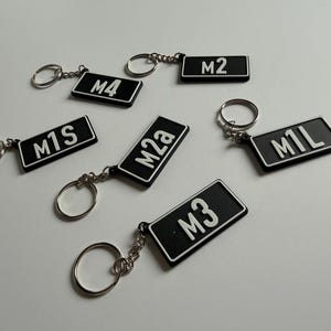 May include: A collection of six rectangular black keychains with white borders and white lettering. Each keychain features a unique alphanumeric code: M4, M2, M1S, M2a, M1L, and M3. Each piece is attached to a circular silver metal split ring.