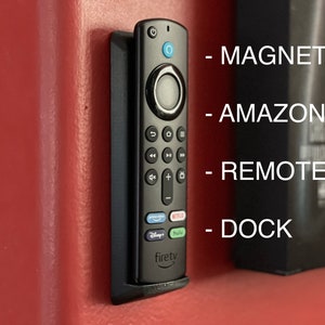 Puede incluir: Un soporte magnético negro para un mando a distancia de Amazon Fire TV. El soporte está montado en una pared roja. El texto "MAGNETIC", "AMAZON", "REMOTE" y "DOCK" es visible en la imagen.
