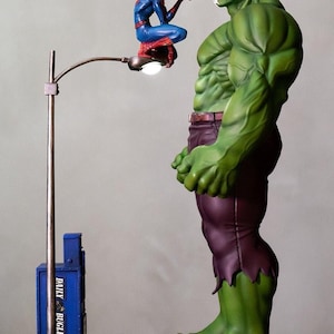 Op de afbeelding: Een verzamelbeeldje met een groene gespierde figuur in een paarse broek die naast een lantaarnpaal staat. Een rood-blauw gekostumeerde figuur zit op de lamp en schiet een wit web naar de groene figuur. Een blauwe Daily Bugle-doos is aanwezig.