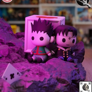 Puede incluir: Dos figuras de estilo anime de personajes con pelo puntiagudo y atuendos distintivos, sentados sobre una superficie rocosa p&uacute;rpura. Una carta de juego con un s&iacute;mbolo de tr&eacute;bol negro est&aacute; en primer plano. Las figuras est&aacute;n junto a una caja blanca.