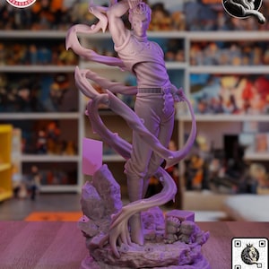 Puede incluir: Figura de color p&uacute;rpura claro de un personaje masculino en una pose din&aacute;mica, con elementos fluidos en forma de cinta. La figura se encuentra sobre una base rocosa con peque&ntilde;as figuras. El fondo est&aacute; borroso, con estanter&iacute;as y otras figuras.