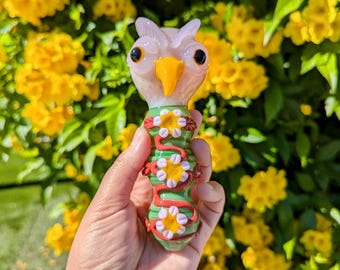 May include: A glass pipe shaped like an owl with a green and red body decorated with white and yellow flowers. The owl has a pink face with yellow beak and black eyes.