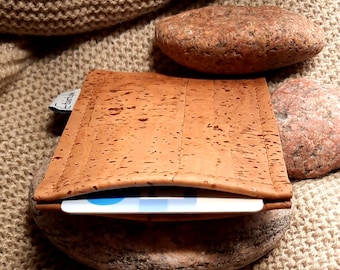 Puede incluir: Una cartera de corcho marrón claro con una tarjeta parcialmente visible. La cartera tiene una superficie texturizada y descansa sobre una piedra. El fondo es de tejido de punto.
