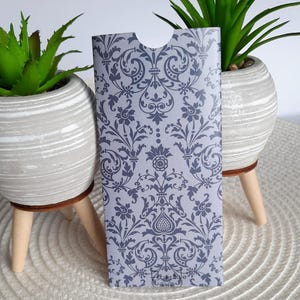 Puede incluir: Un sobre gris con un patrón de damasco negro. El sobre tiene una ranura cortada en la parte superior para insertar una tarjeta o una tarjeta de regalo. El sobre tiene la palabra "Lifestyle" impresa en la parte inferior.