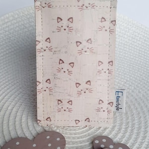 以下が含まれることがあります： 猫の顔が繰り返される模様の、薄いピンク色のピルケース。ケースにはピルのブリスターパックが入っています。2つの木製のハートの装飾が前景にあります。タグには「Etwistyle」と書かれています。