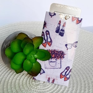 Könnte beinhalten: Ein dekorativer Halter mit einem Aufdruck von Schuhen, Lippenstift und Brillen, der eine Blisterpackung mit Pillen enthält. Eine grüne Sukkulente befindet sich in einem grauen, steinartigen Behälter. Auf dem Halter steht das Wort "Flowers".