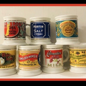 May include: A collection of seven vintage ceramic mugs with gold trim. Each mug features a different brand label, including "Il BROS Java Mocha Coffee," "Morton Salt," and "Whitman's Sampler." The mugs are arranged on a white shelf.