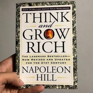 Może przedstawiać: Książka "Think and Grow Rich" autorstwa Napoleona Hilla. Okładka z czarnym tekstem na białym tle i małym obrazkiem płomienia. Krawędzie książki zdobi wzór z banknotami. Książka jest trzymana w dłoni.