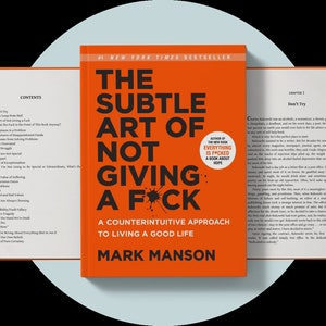May include: An open book with an orange cover titled "The Subtle Art of Not Giving a F*ck" by Mark Manson. The book is a New York Times bestseller and has a counterintuitive approach to living a good life.
